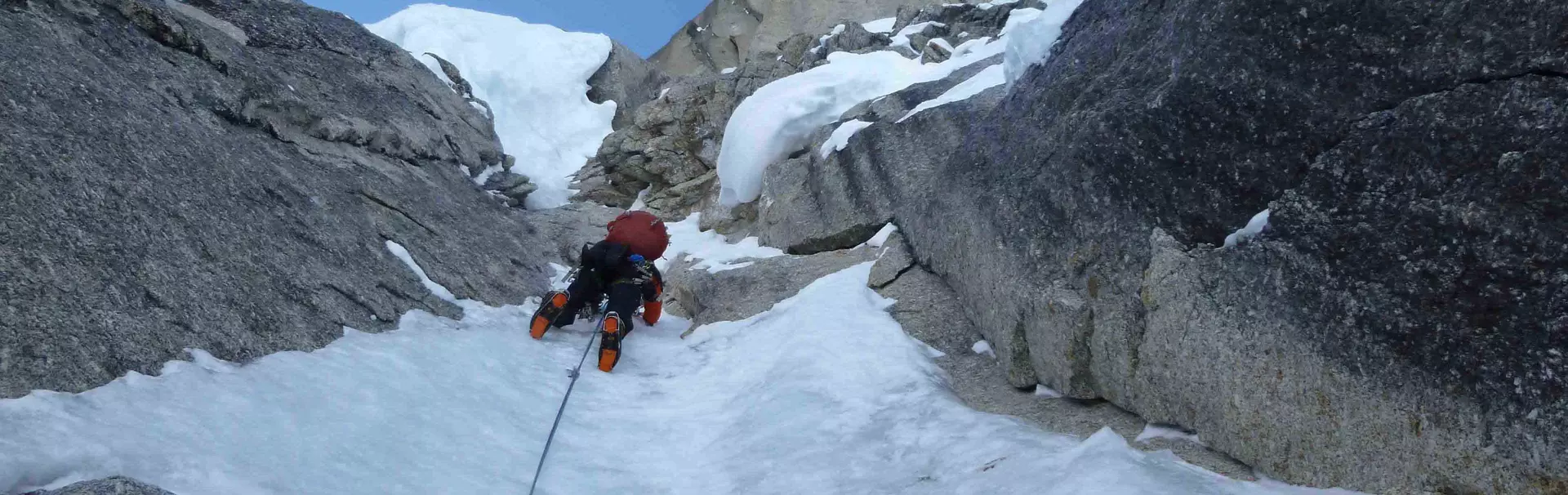 Een steile, ruige berg, bedekt met sneeuw en ijs, rijst schril op tegen een gedeeltelijk bewolkte blauwe lucht, terwijl het zonlicht de top van achteren verlicht.